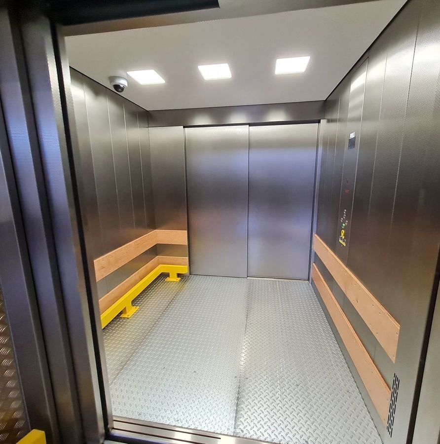 Innenraum eines Aufzugs mit Metallwänden, gelber Sitzbank und geschlossener Doppeltür